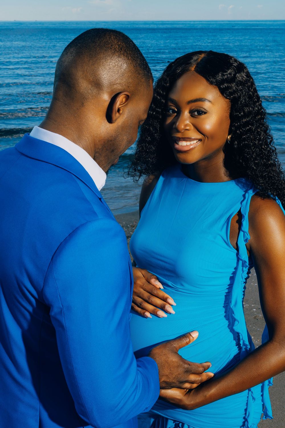 A couple in vibrant blue outfits shares a tender moment on the beach during a maternity photoshoot by the ocean.