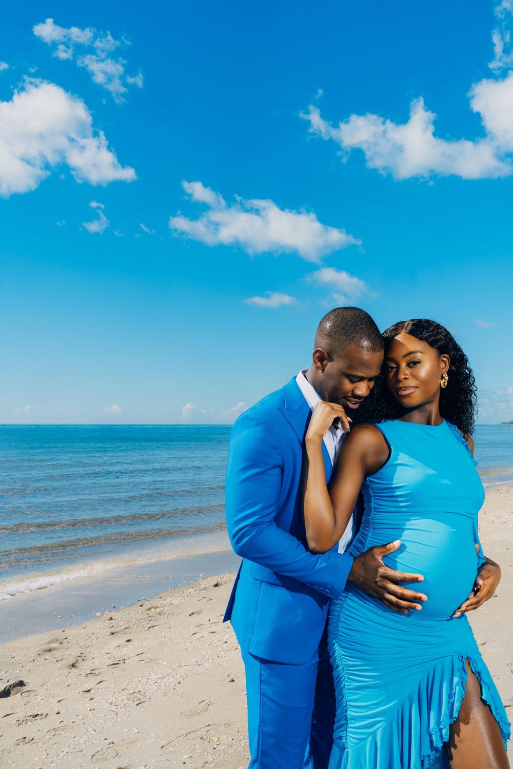 A couple in matching blue outfits embrace lovingly on a sunny beach with turquoise waters and white sand.