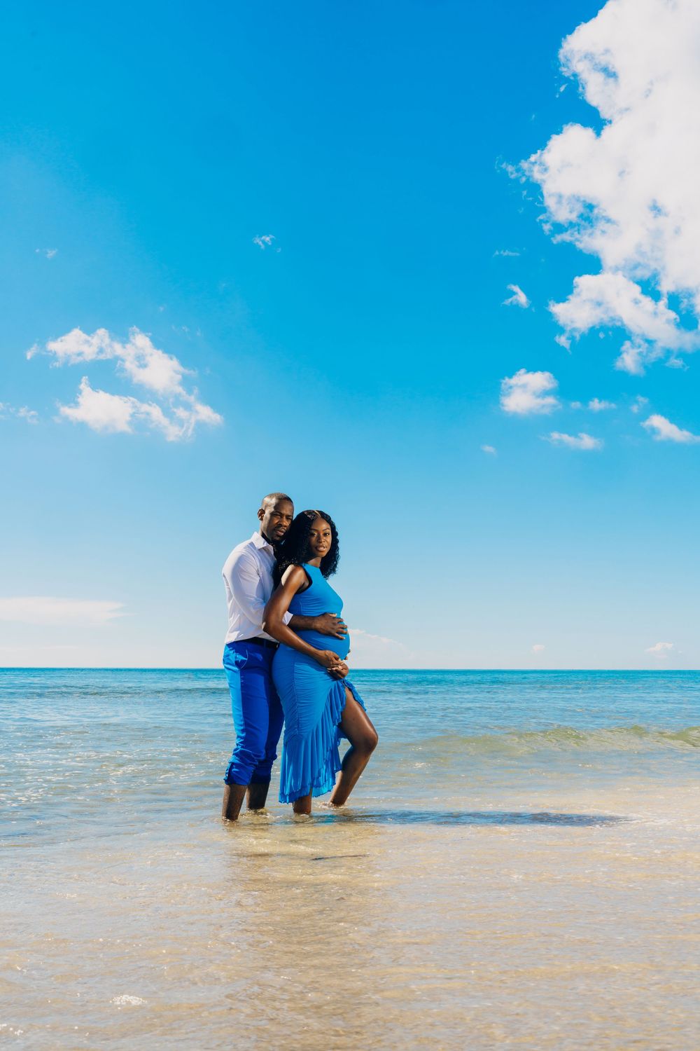 Couple standing in shallow ocean water on sunny beach under bright blue sky with white fluffy clouds.