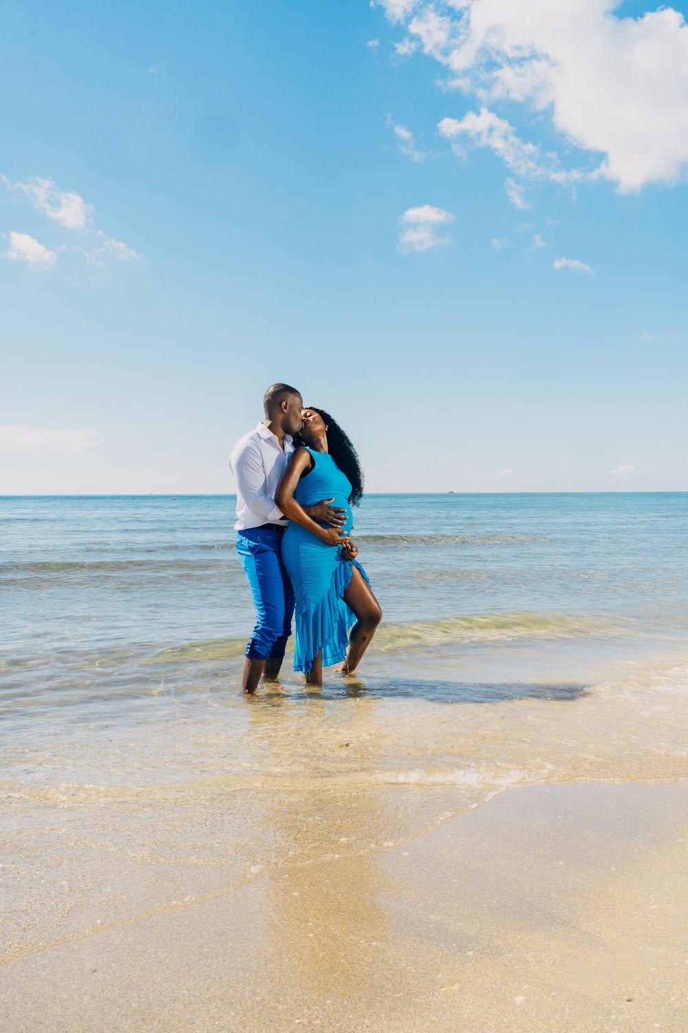 A couple embraces while standing in shallow ocean water on a sunny beach with blue skies overhead.
