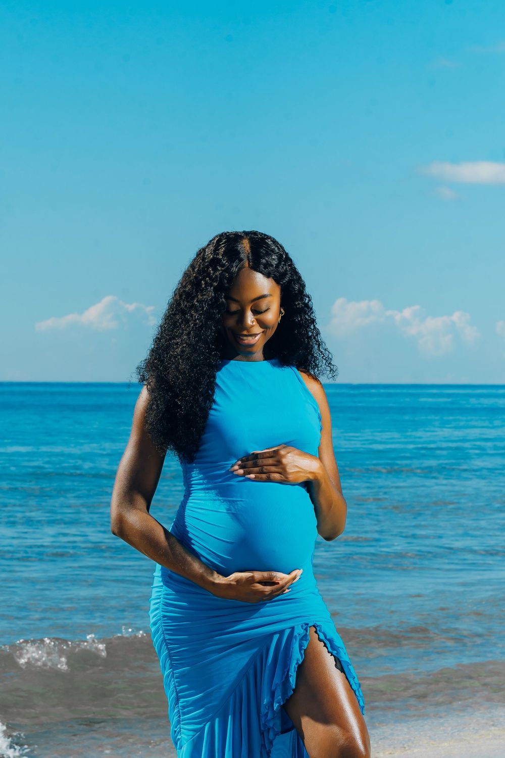 A pregnant woman in a bright blue dress poses gracefully on a tropical beach with turquoise ocean waters and blue sky.