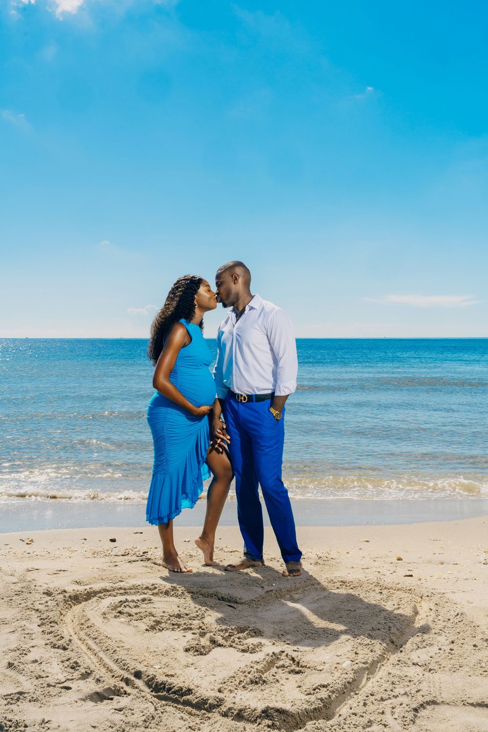Couple in blue outfits share a romantic moment on the beach while standing in a heart drawn in the sand.