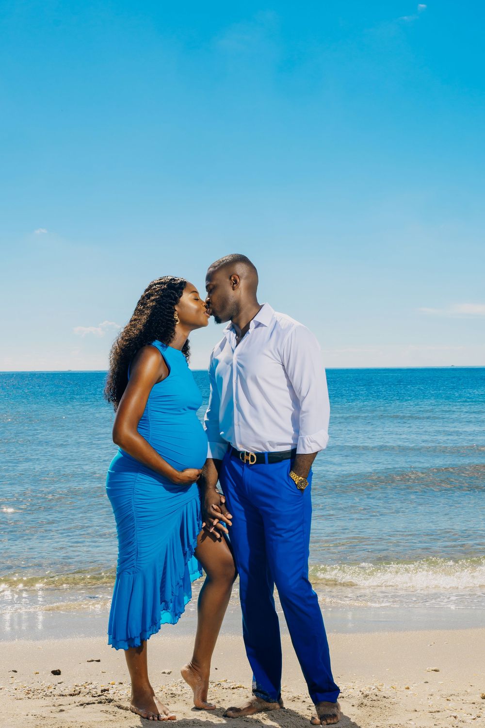 Couple in coordinating blue outfits share a romantic moment on a sunny beach with ocean waves in background.