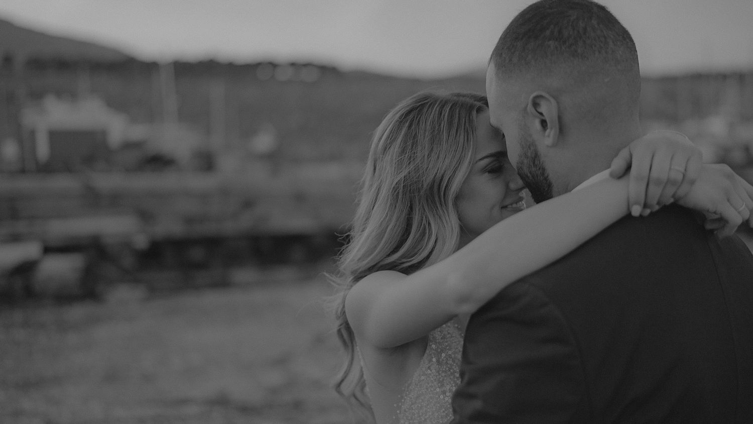 A couple embraces in a tender moment against a blurred outdoor landscape in black and white photography.