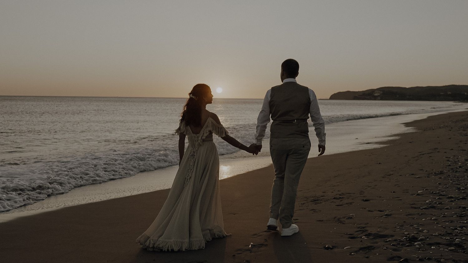 A couple holds hands while walking along a misty beach at sunset with ocean waves visible in the background.