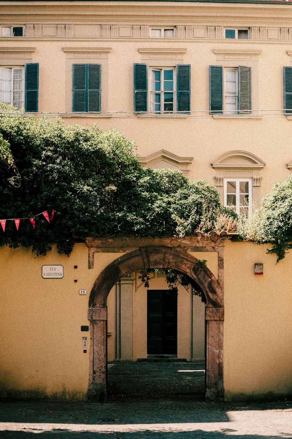 Historic Italian building with arched entrance, green shutters, and climbing vines under warm sunlight.