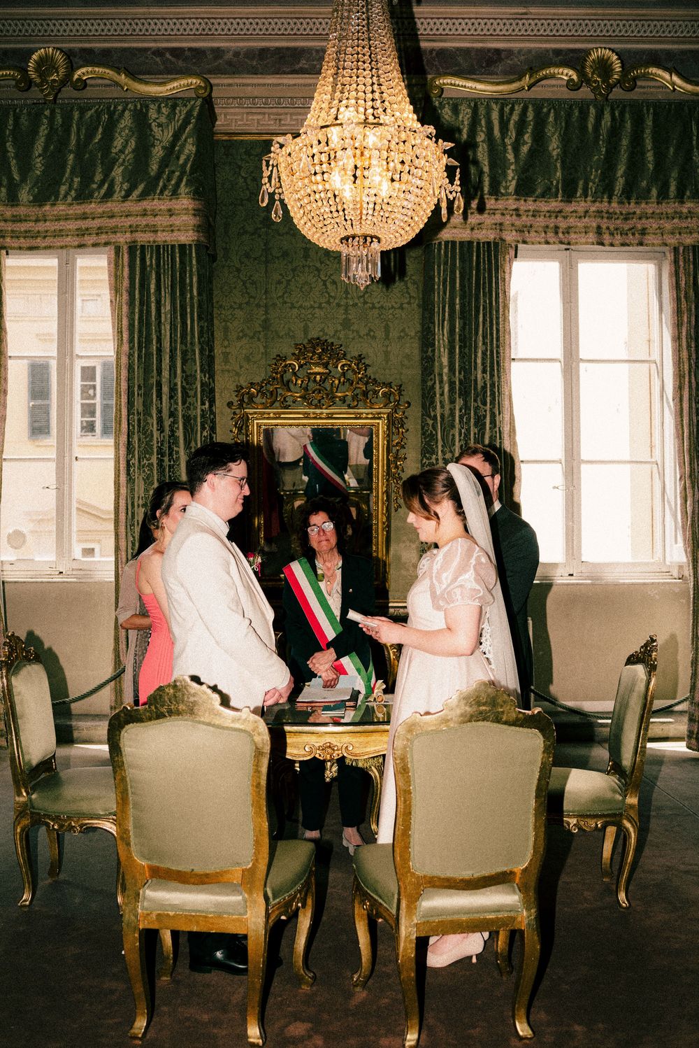 Elegant wedding ceremony in ornate green room with chandelier, couple exchanging vows before officiant wearing Italian sash.