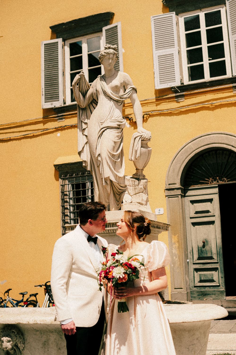 Bride and groom pose before a marble statue and yellow Italian building, bride holding a vibrant bouquet.