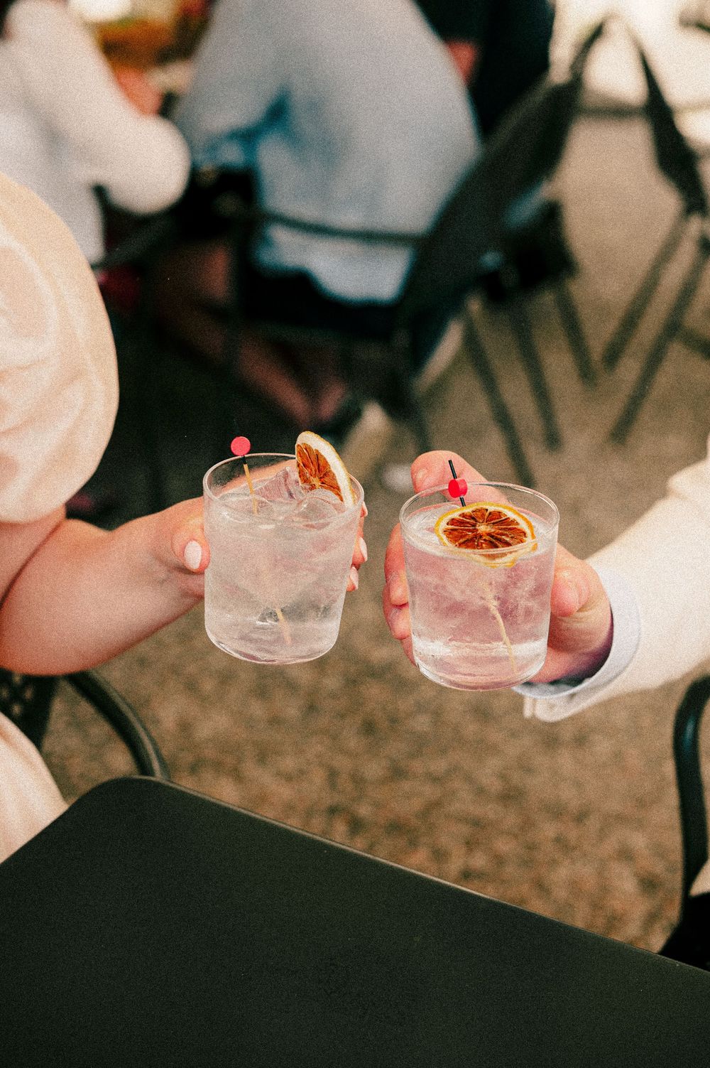 Two people toasting with iced cocktails garnished with dried orange slices and pink picks at an outdoor venue.
