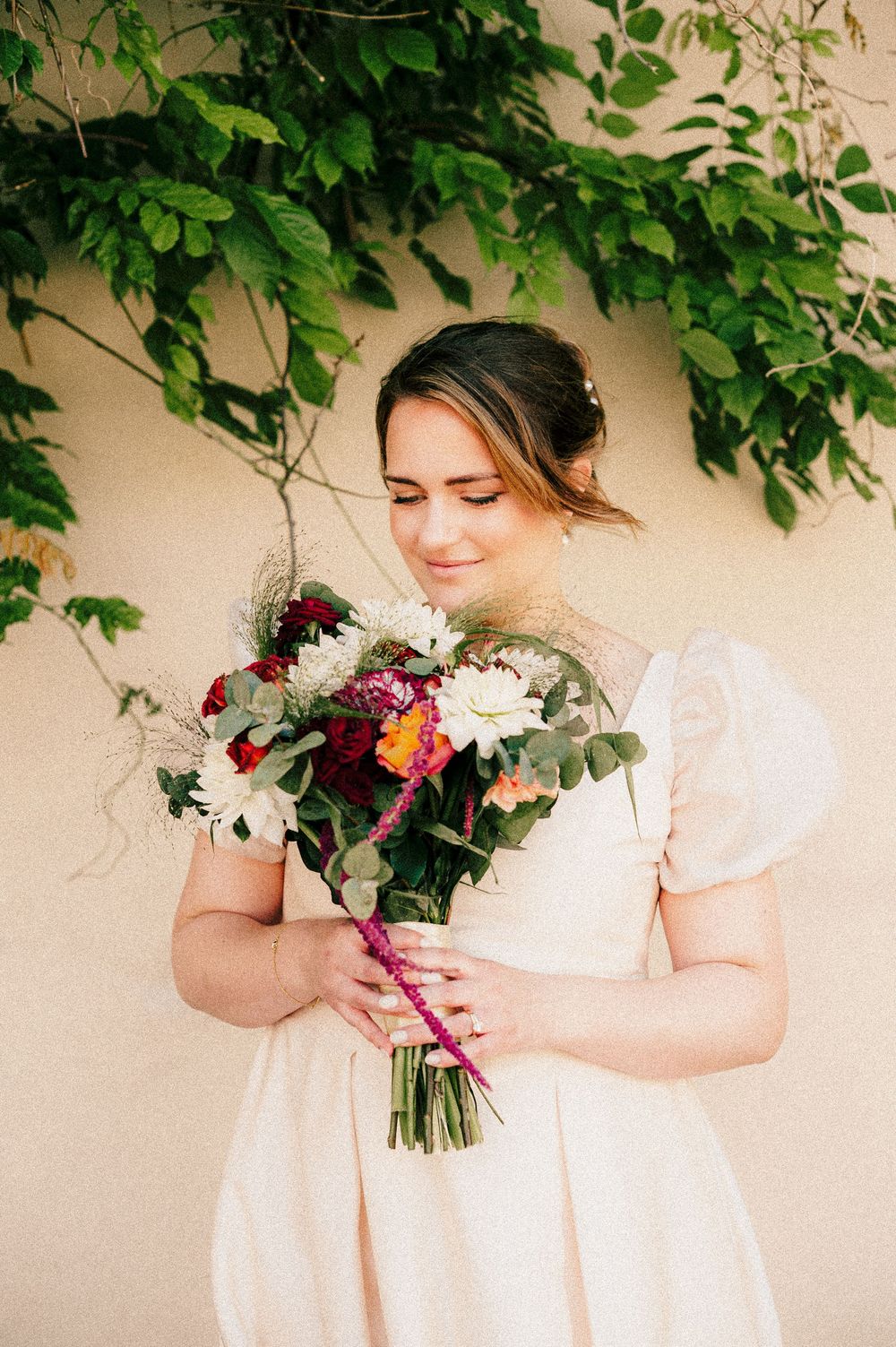 Bride in white dress holding colorful wedding bouquet with dahlias and greenery, smiling softly outdoors.