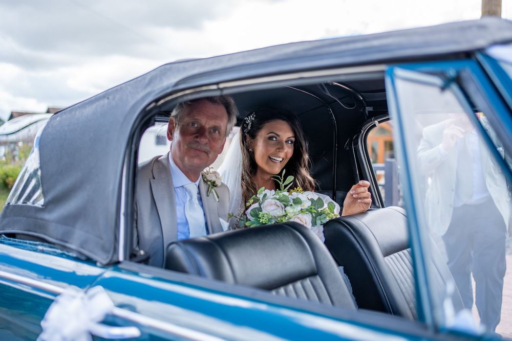 Happy passengers wave from vintage blue classic cars decorated with floral arrangements at a celebratory event.