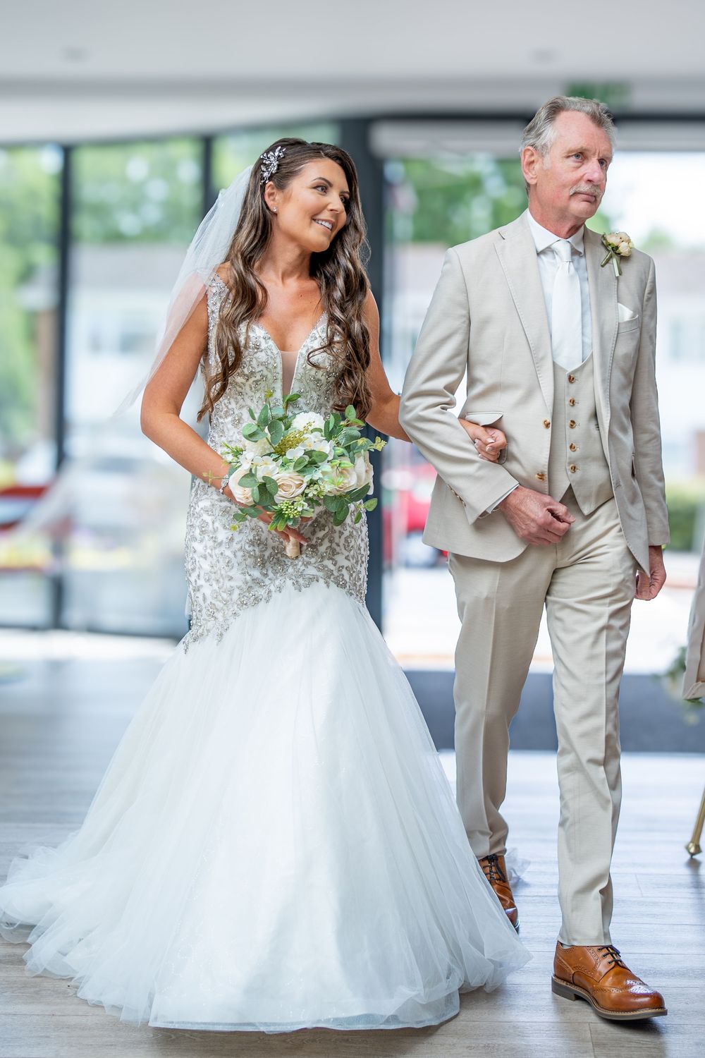 A bride in a sparkling white mermaid gown walks arm-in-arm with a man wearing a light beige three-piece suit.