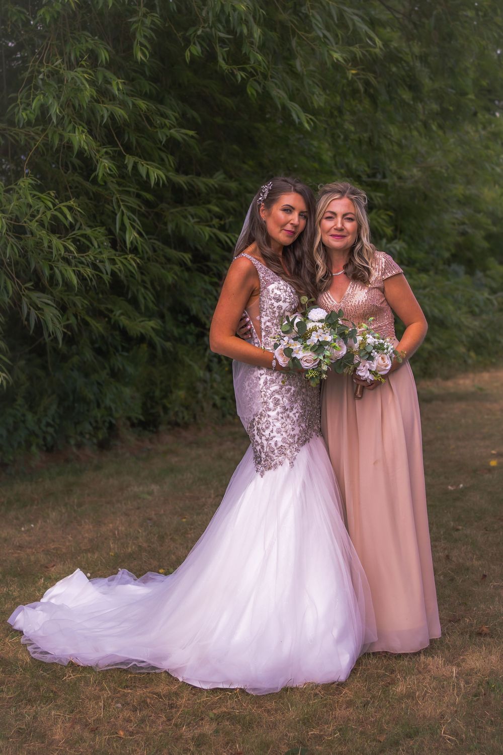 Two people pose outdoors in formal attire, wearing a white mermaid wedding gown and blush pink bridesmaid dress with bouquets.