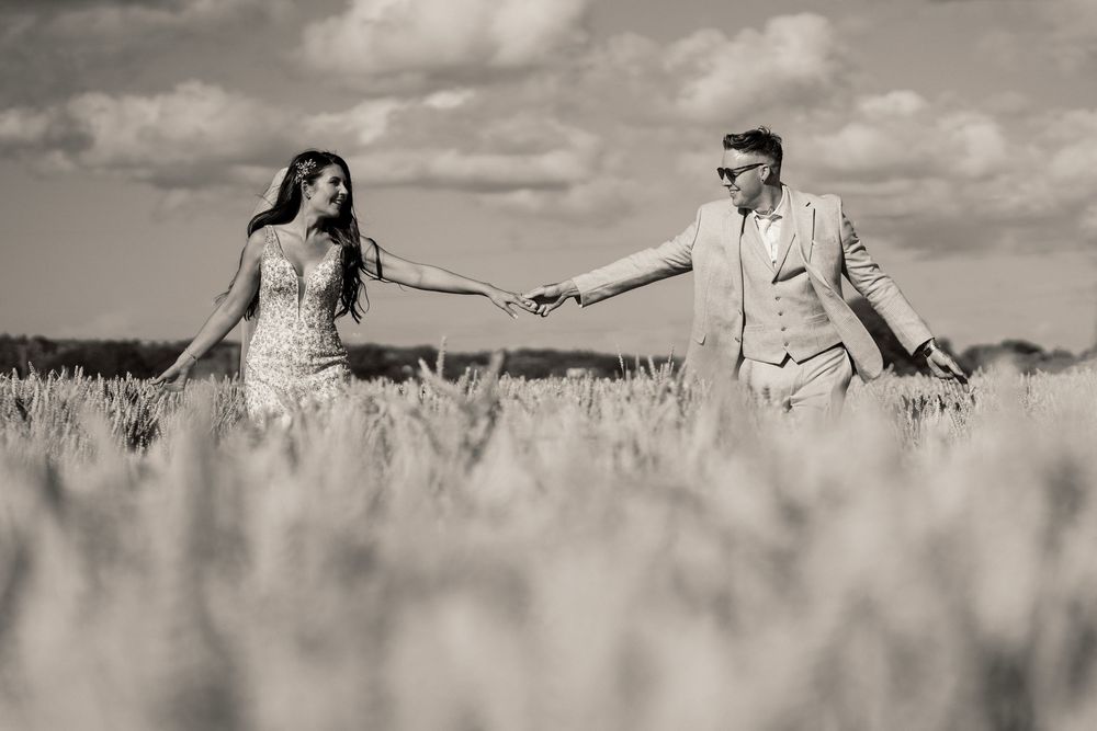 Black and white romantic photo of a couple holding hands while walking through a wheat field on an overcast day.