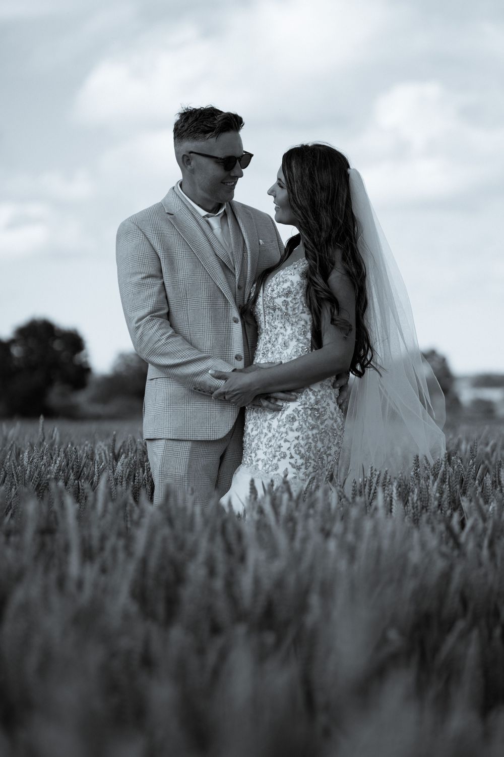A romantic black and white wedding photo of newlyweds embracing in a wheat field on an overcast day.