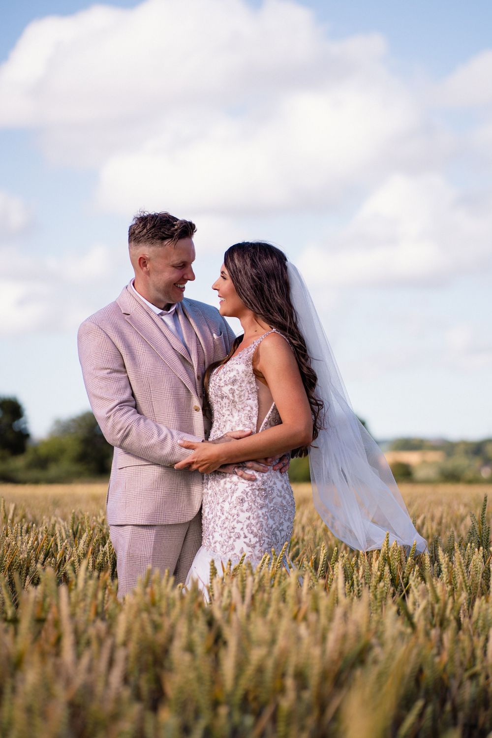 Romantic wedding couple embraces in golden wheat field under blue sky with white clouds and flowing veil.