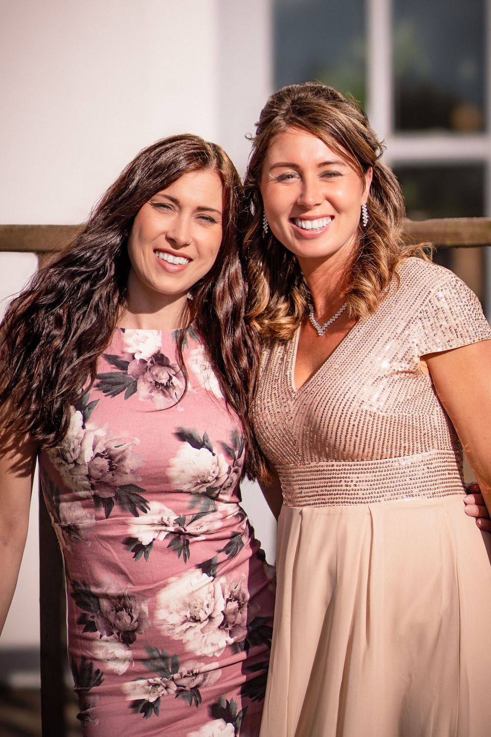 Two women wearing pink dresses smile together in an indoor portrait with natural lighting.