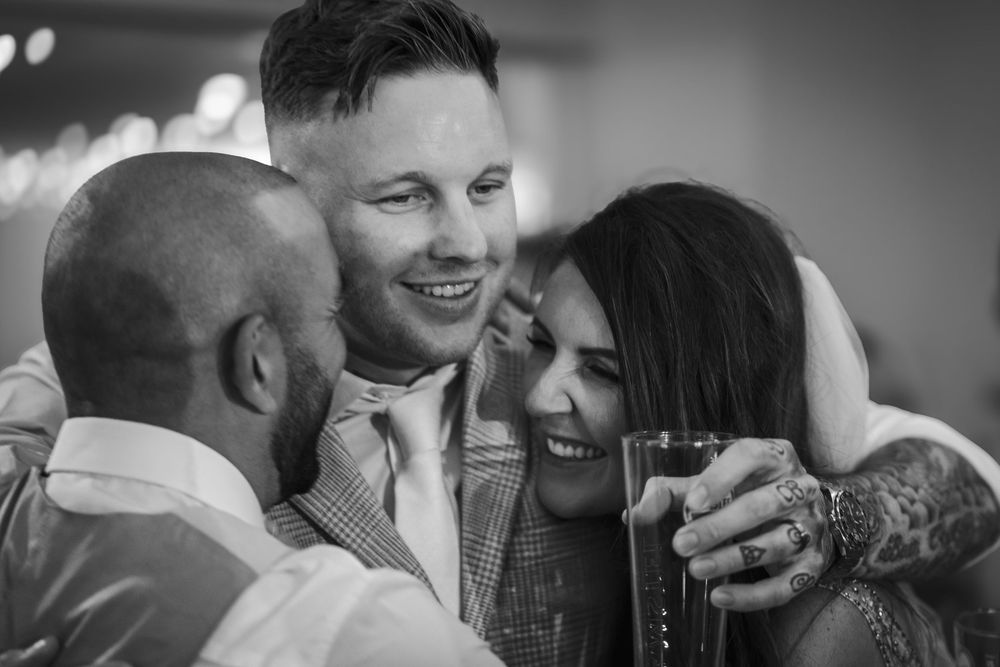 A candid black and white photograph capturing three friends sharing a joyful, intimate moment at a celebration.