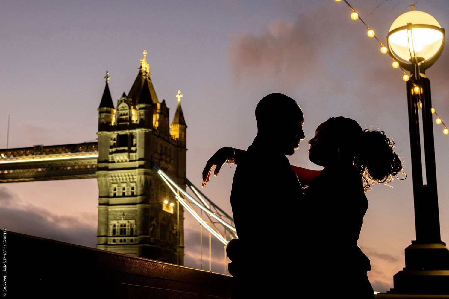 Silhouetted romantic couple against dramatic sunset with illuminated Tower Bridge and vintage street lamp in London.