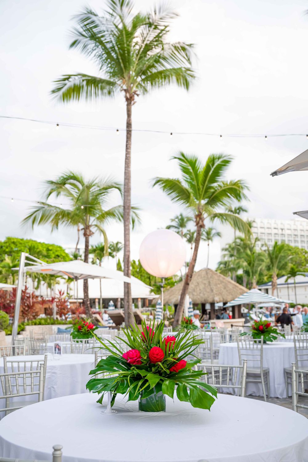 Outdoor reception setup with white tables, palm trees, and tropical floral arrangements.