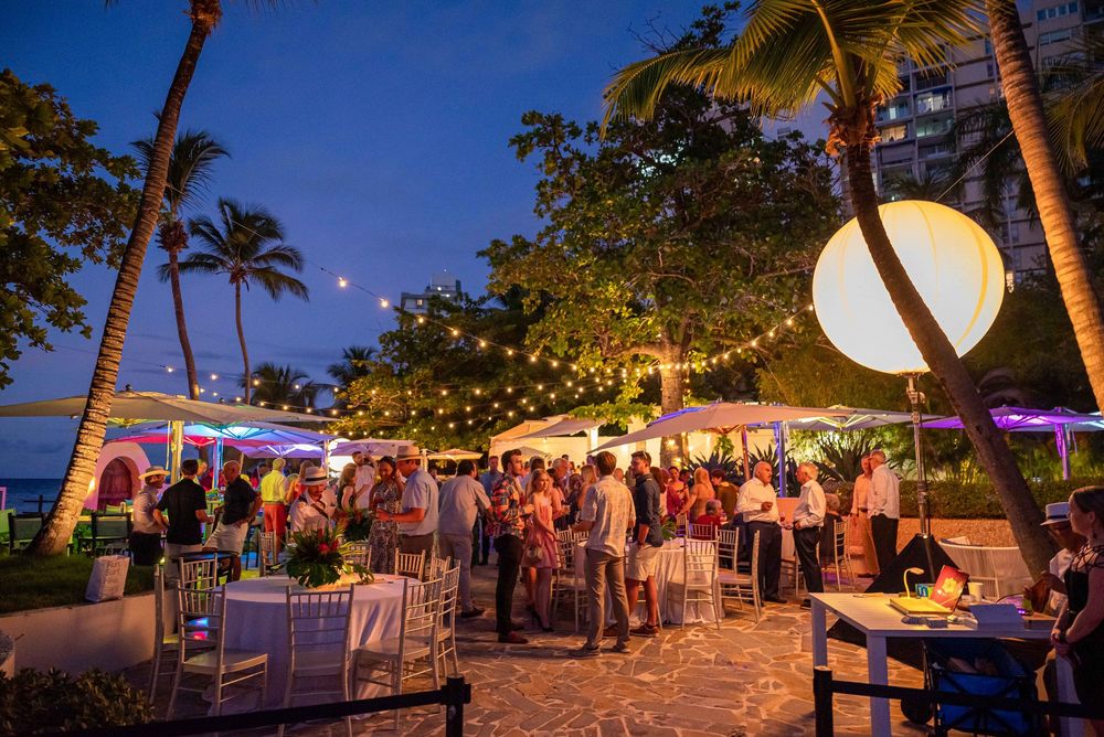 Evening outdoor reception with string lights, palm trees and guests mingling on a beachfront patio.