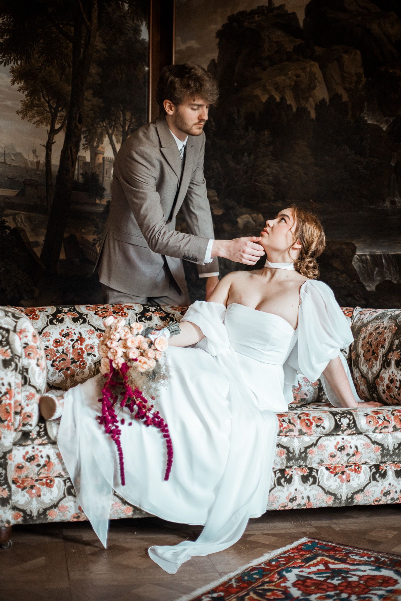 A romantic couple in wedding attire share an intimate moment on a floral patterned sofa in a dark moody setting.