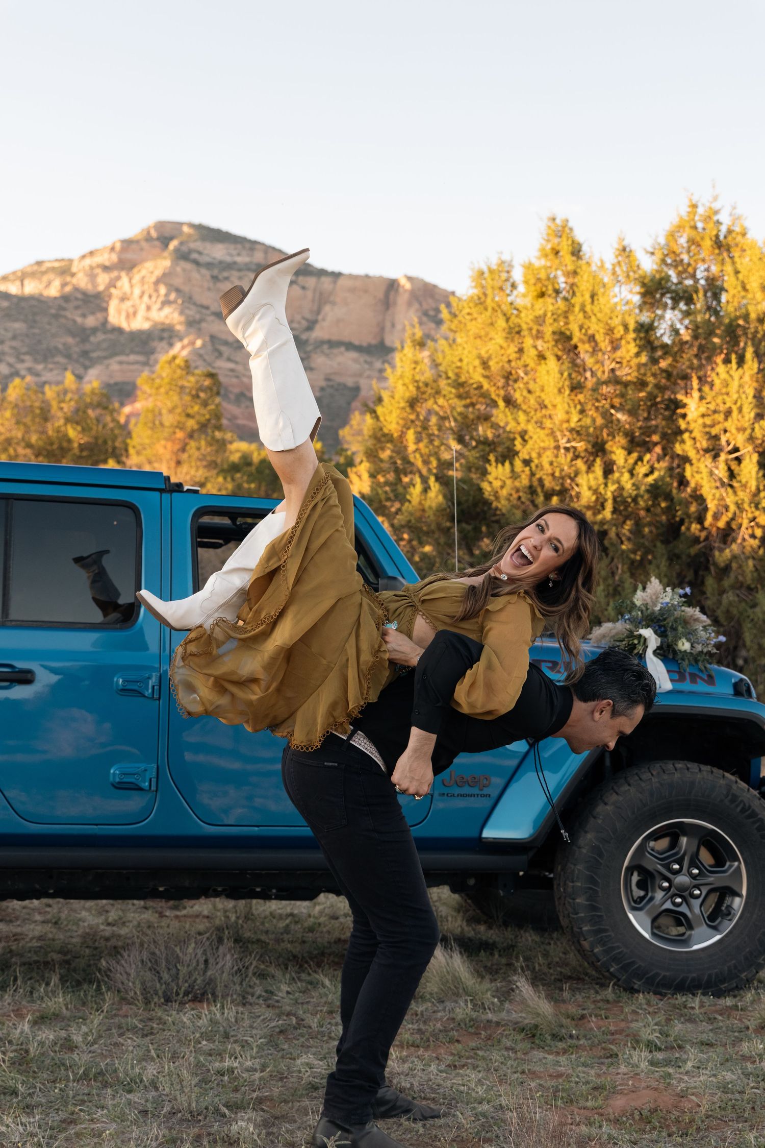 A dancer in a brown jacket performs a high kick pose in front of a blue Jeep with autumn trees and mountains in the background.