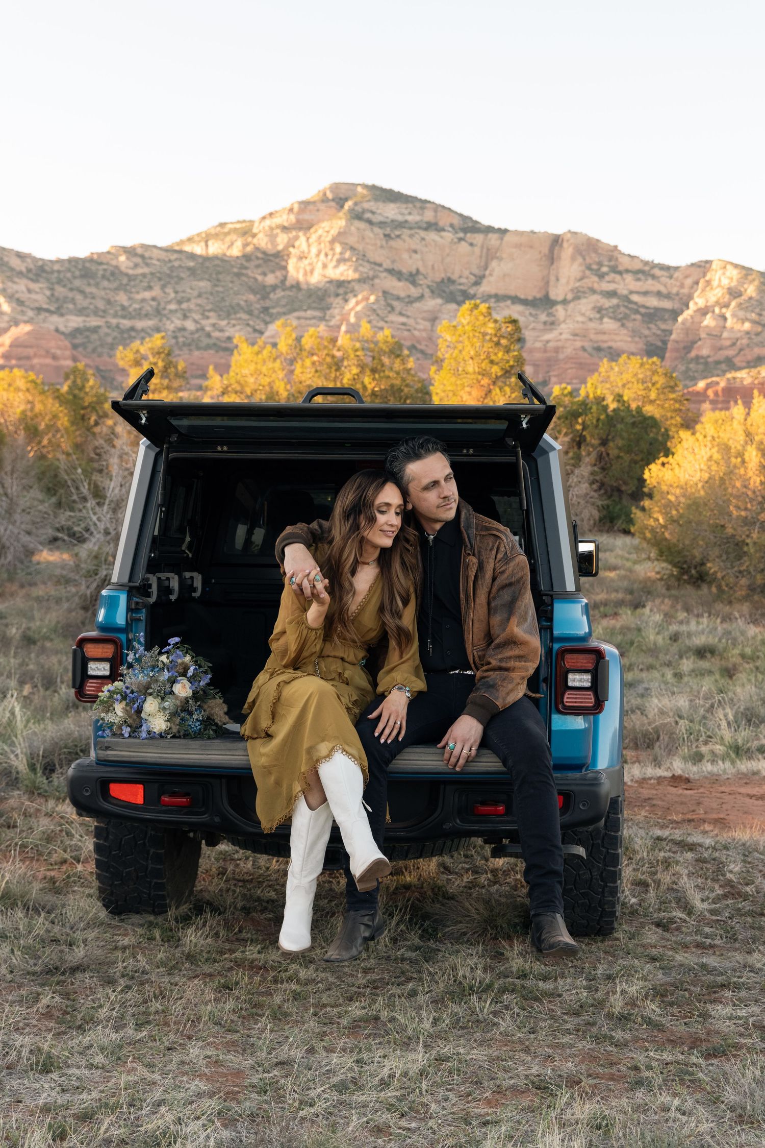 A couple sits together in the back of a blue Jeep with mountain scenery and fall foliage in the background.