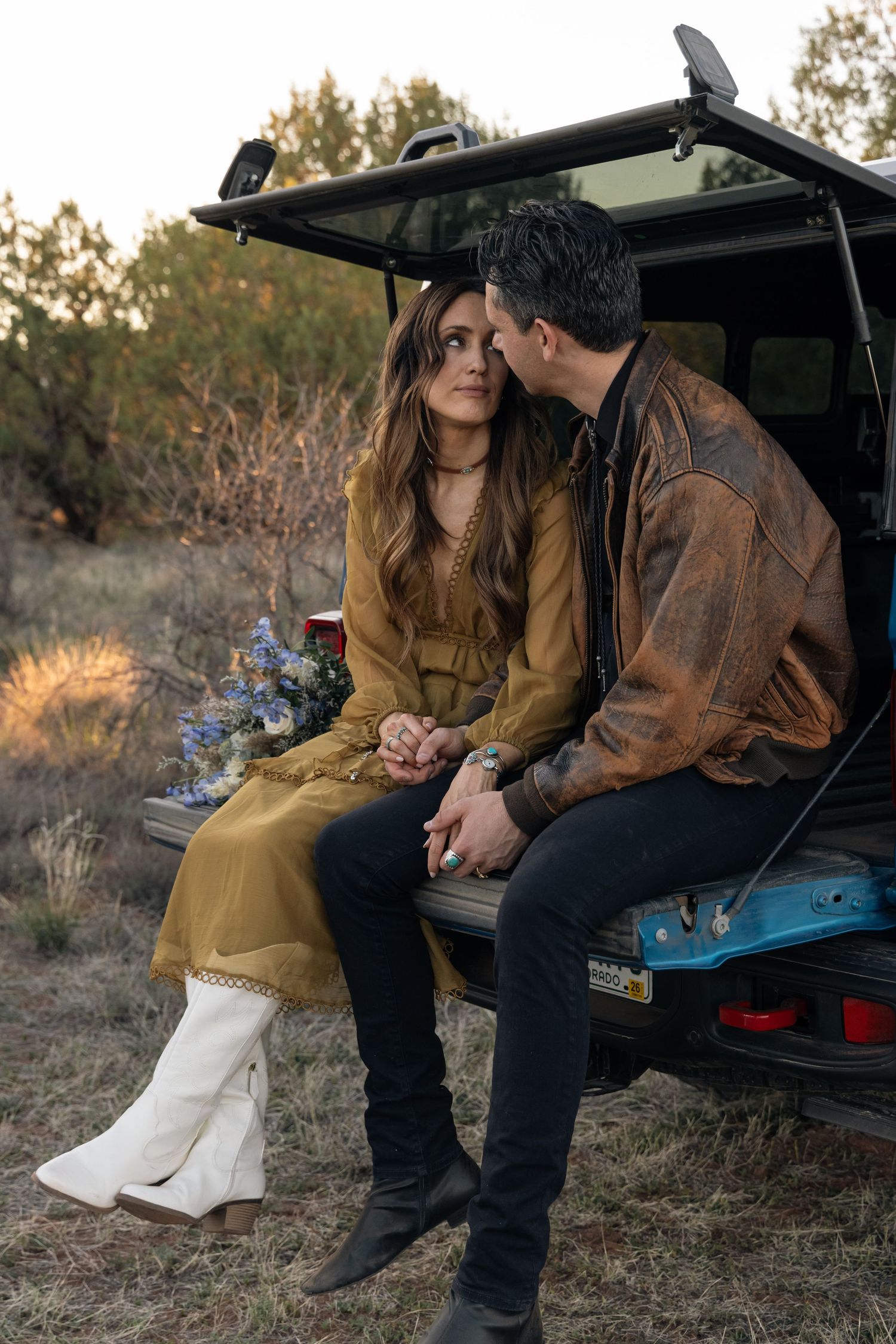 A couple sits together on the tailgate of a vehicle in a desert setting at sunset wearing brown and white boho attire.