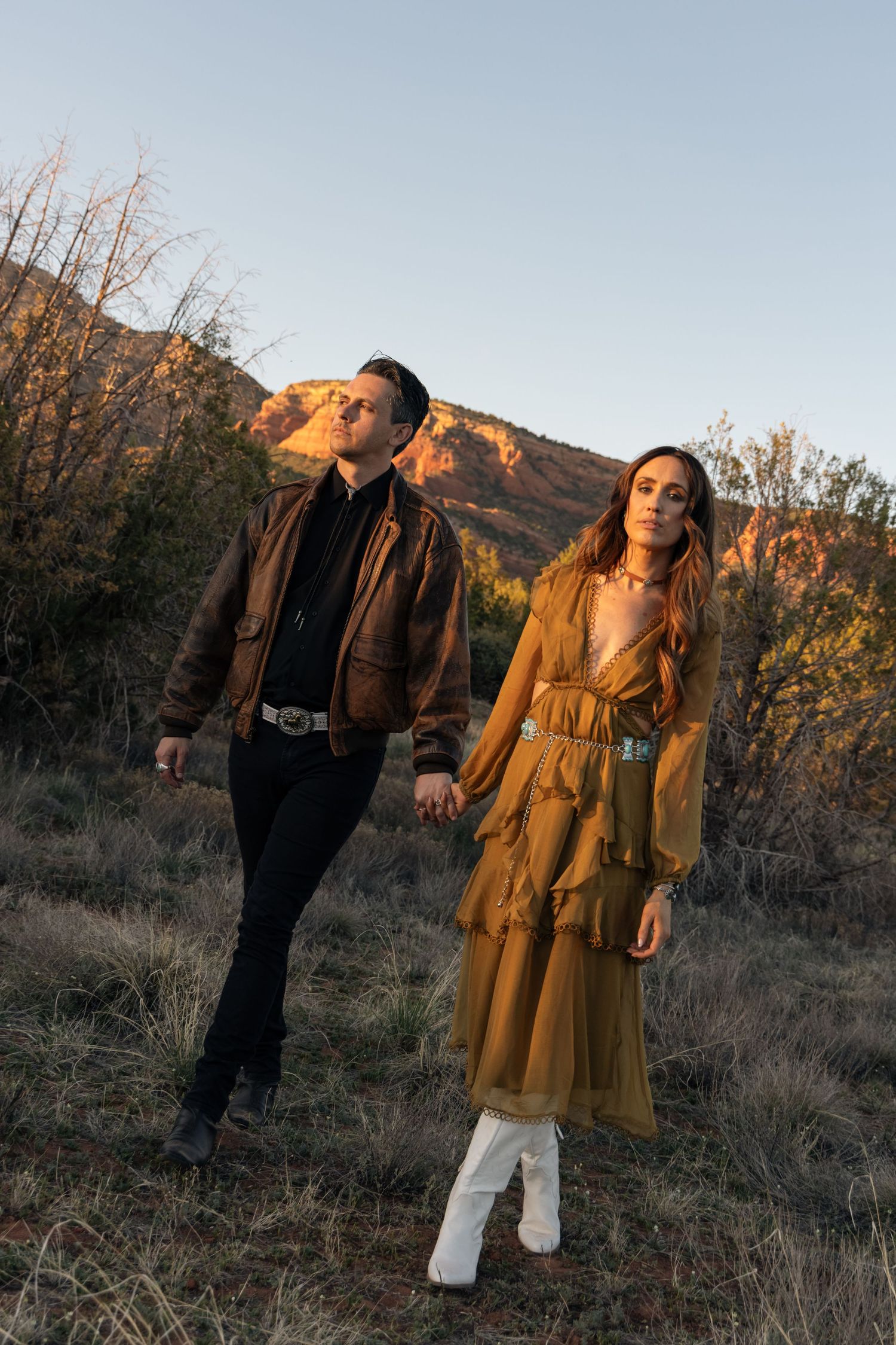 A couple poses outdoors in autumn attire, with a leather jacket and mustard ruffle dress against a mountainous backdrop.