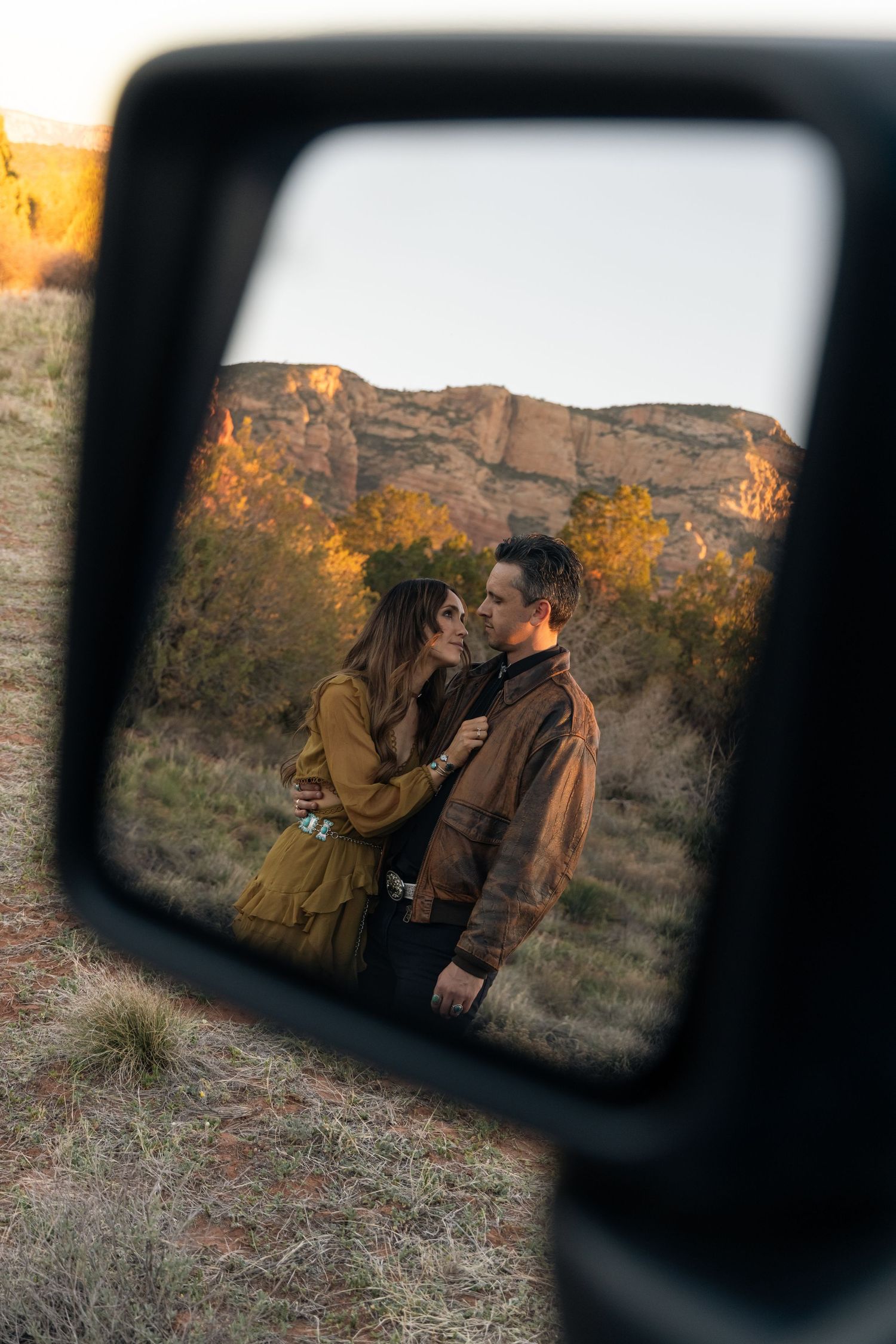 A romantic mountain scene reflected in a car side mirror during sunset shows a couple embracing against an autumn landscape.