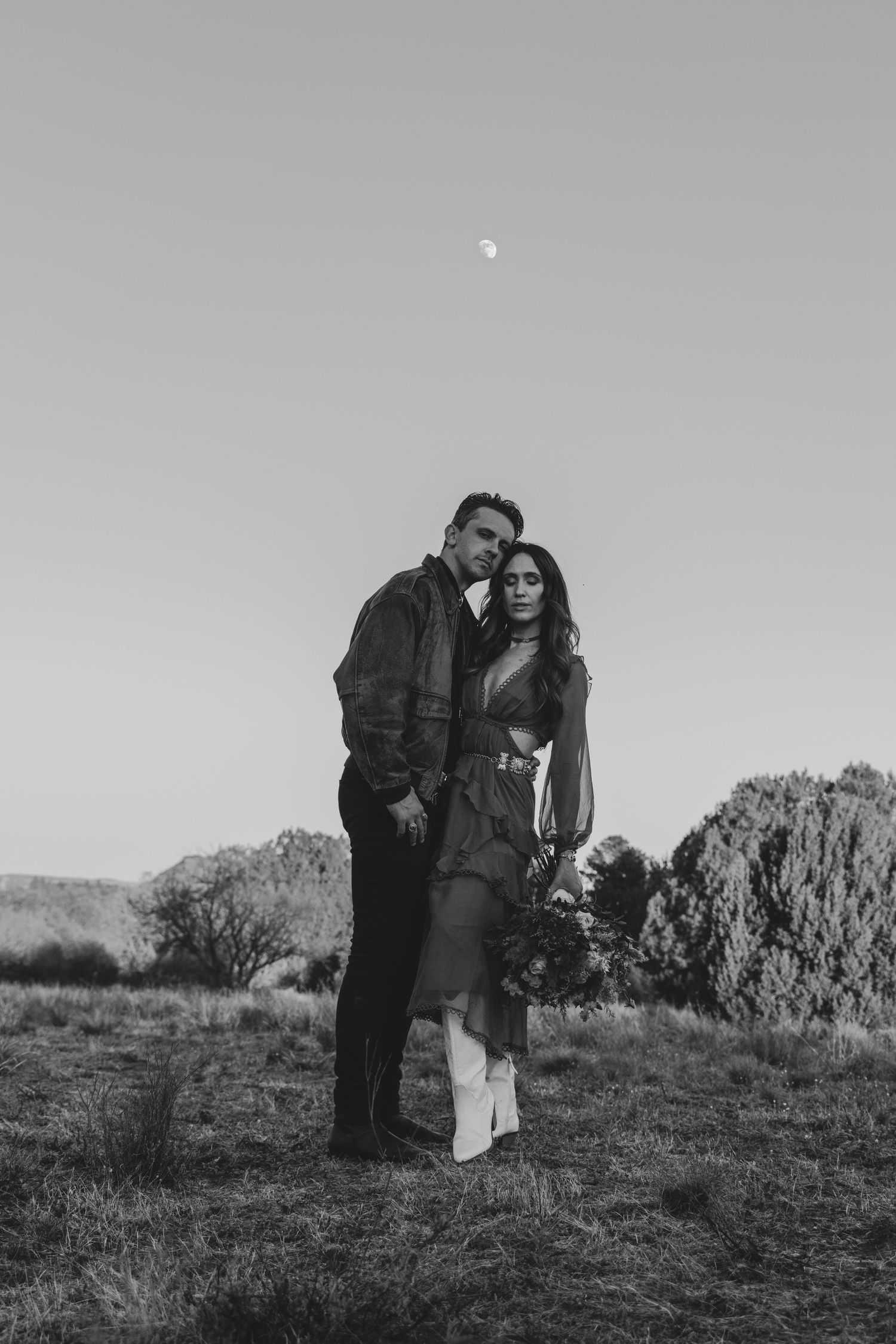 A romantic black and white photo of a couple embracing outdoors under a pale moon in a rocky landscape.