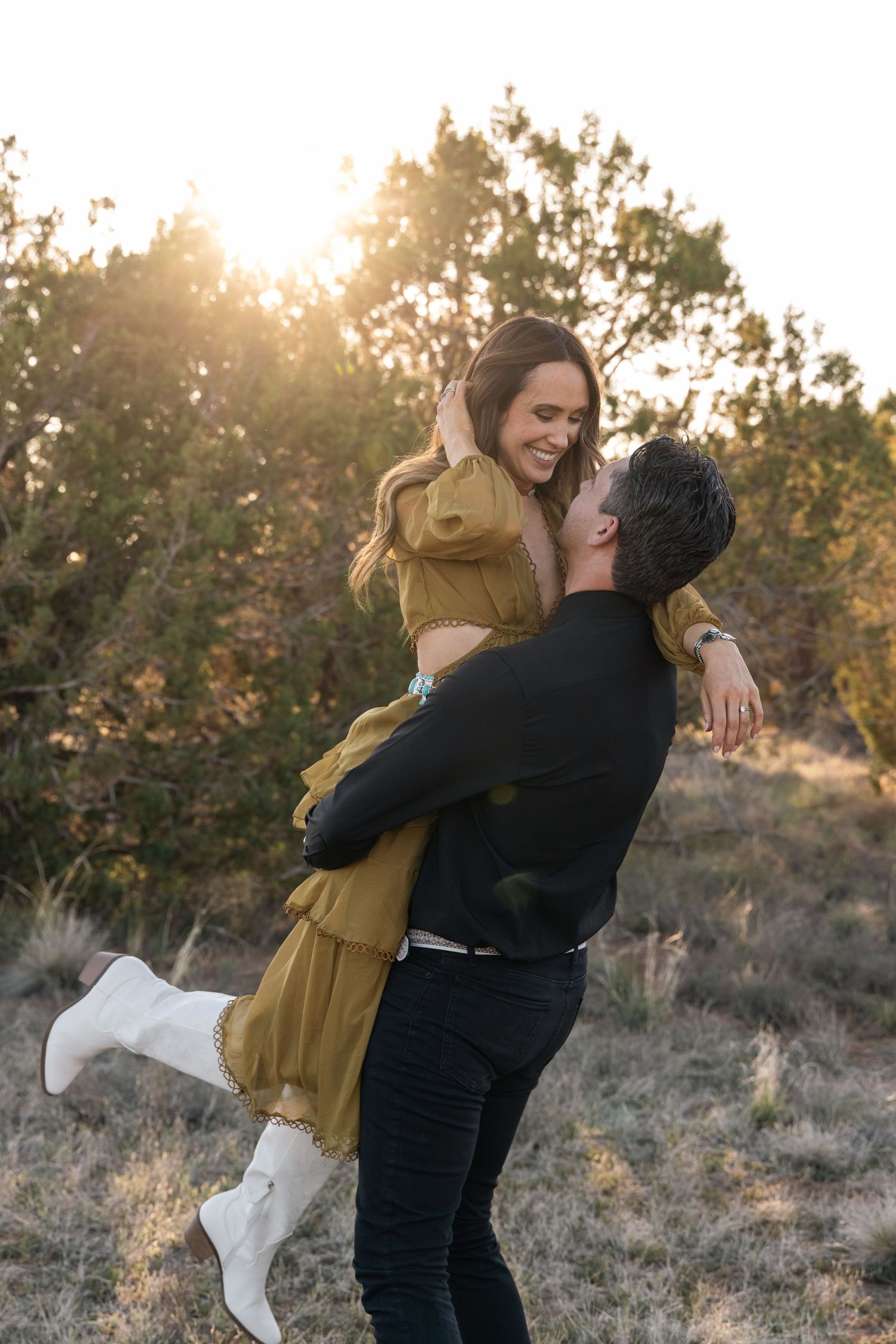 Couple embraces in sunset light with person in mustard dress and white boots being lifted by partner in black clothing.