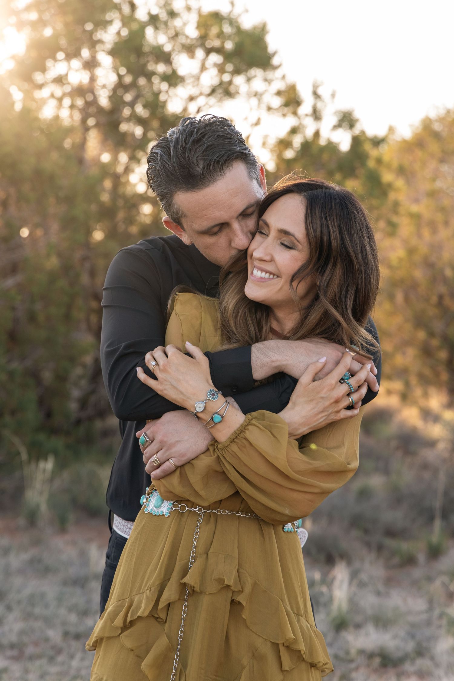 Romantic couple embraces outdoors at sunset as woman in mustard dress smiles while being held from behind.