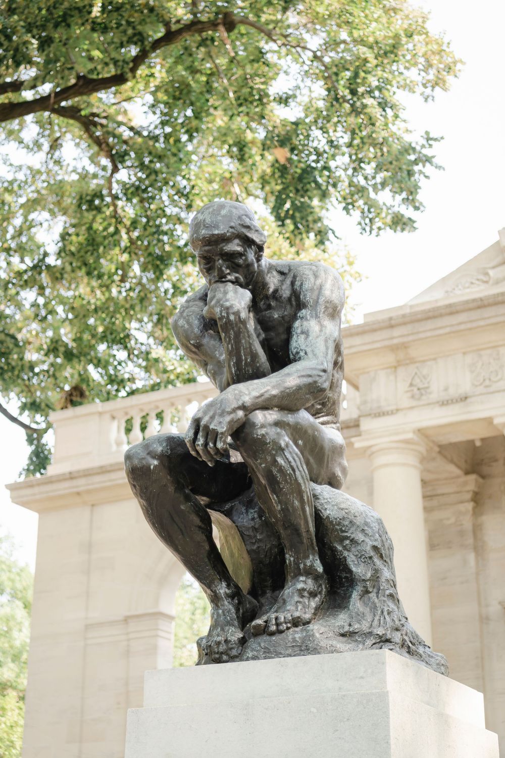 A bronze sculpture of The Thinker by Rodin sits on a pedestal in front of a classical building with trees in background in Philadelphia.