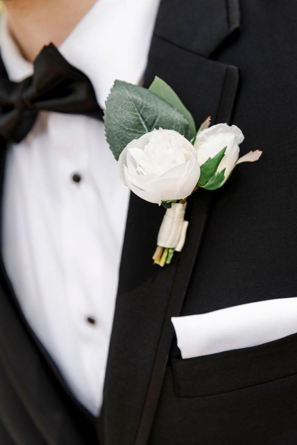 Elegant white rose boutonniere with green leaves pinned to a classic black tuxedo lapel next to white pocket square.