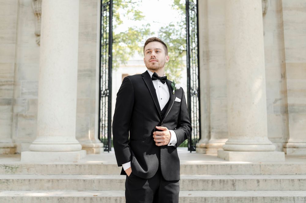 An elegant groom in a classic black tuxedo and bow tie stands between white marble columns on stone steps.