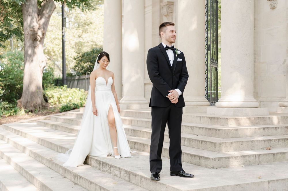 Bride in sleek white dress with slit and groom in classic black tuxedo stand on elegant stone steps of the Rodin Museum for their first look