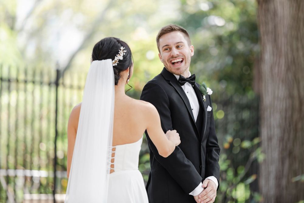 A bride in a white veil and groom in black tuxedo share a joyful first look moment in an outdoor garden setting.