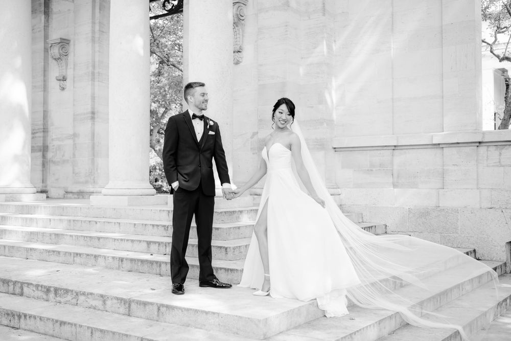 Black and white wedding photo of a couple in formal attire standing on stone steps of a grand building.
