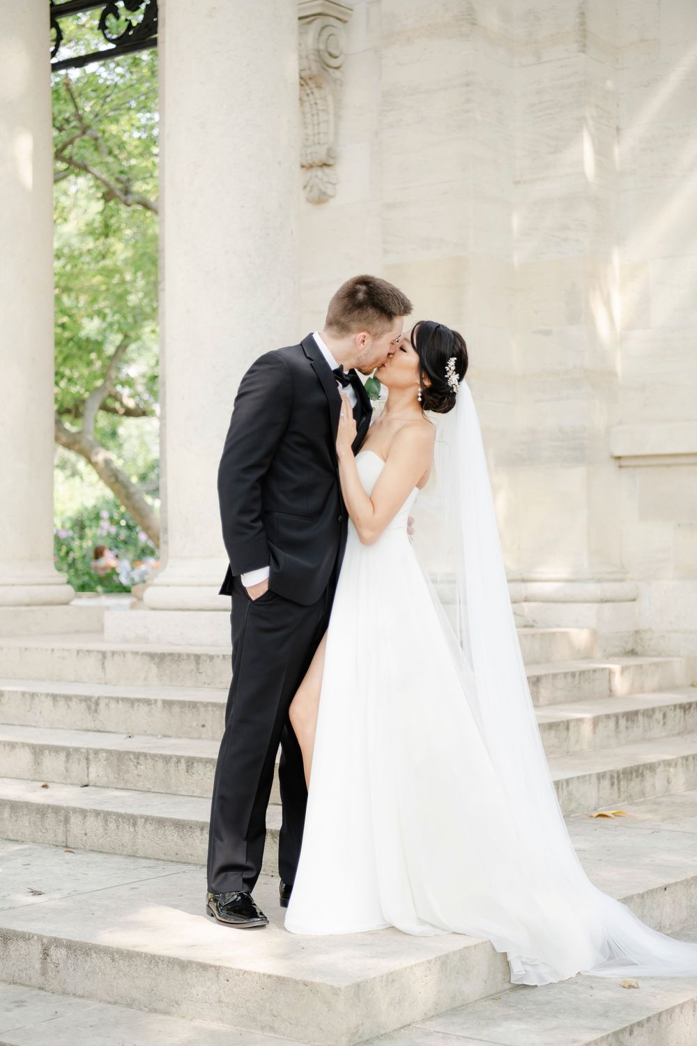A romantic wedding couple stands on white stone steps in an elegant outdoor setting with classical columns and greenery.