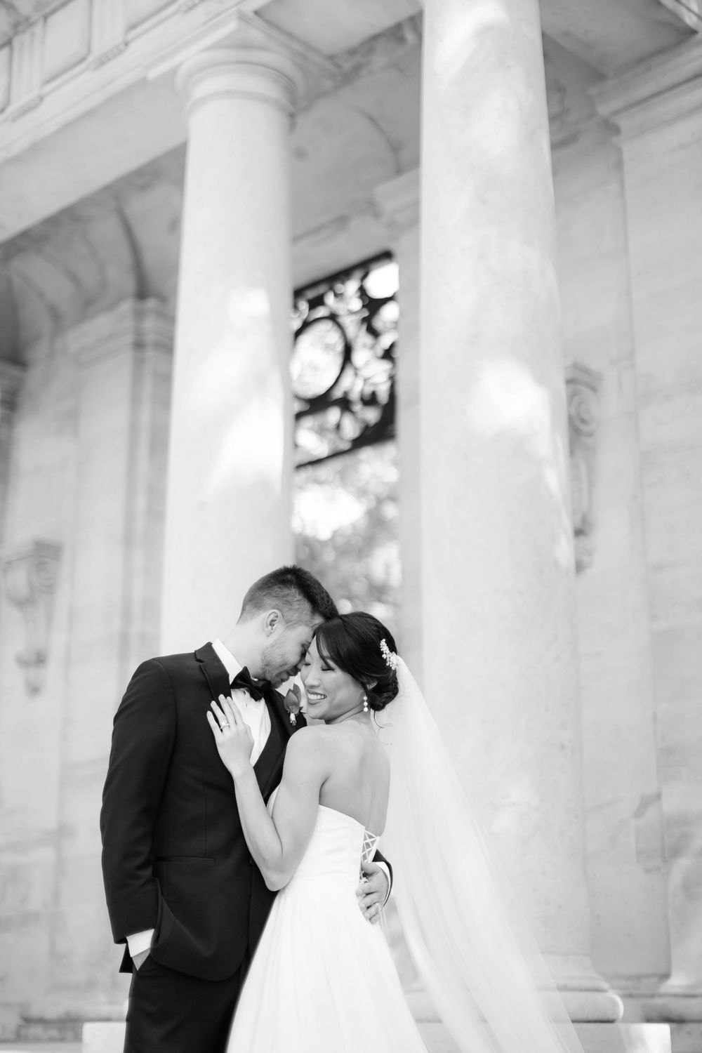 A romantic black and white wedding photo shows a couple embracing between elegant white columns of a classic building.