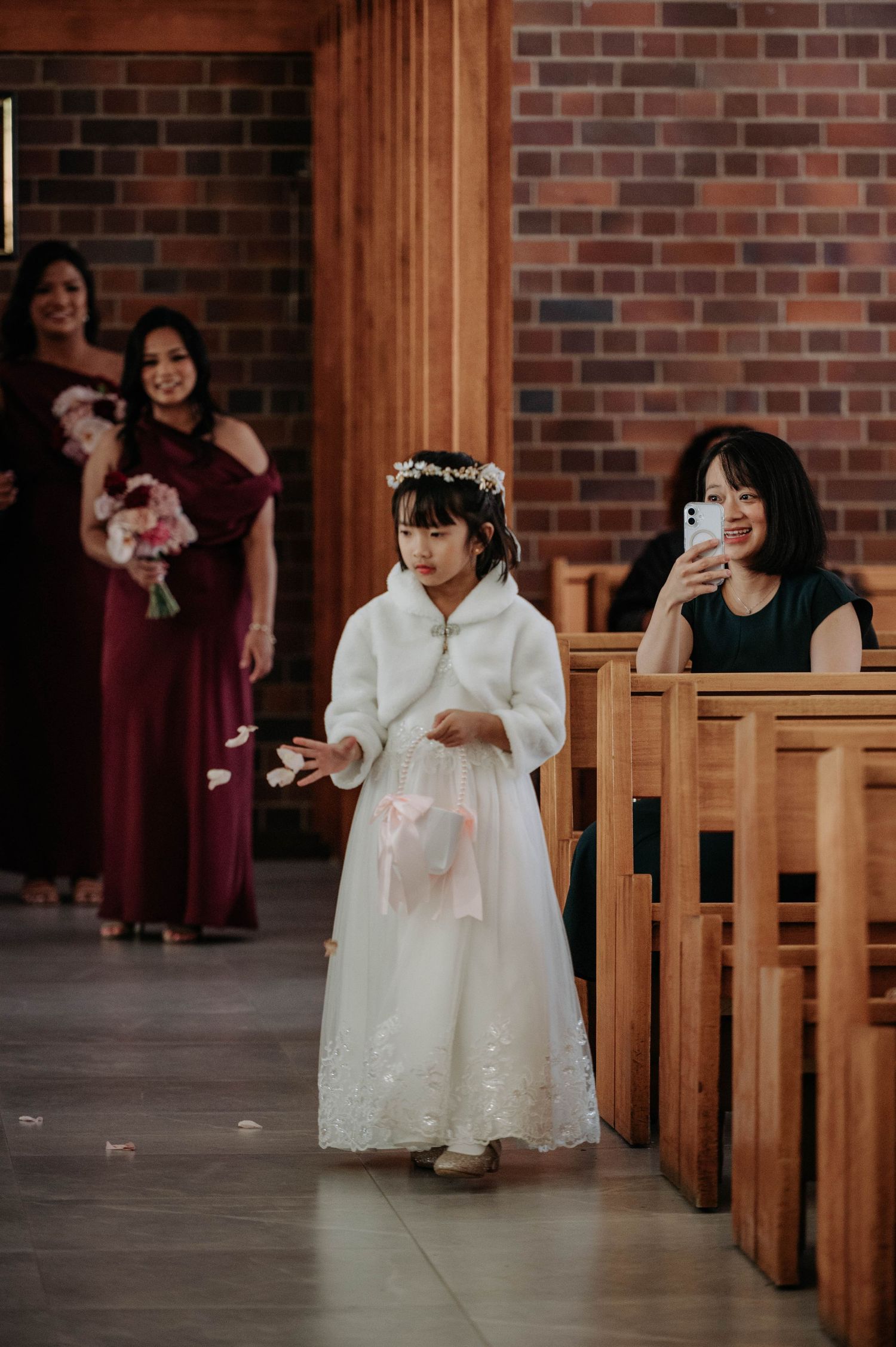 A young flower girl in white dress walks down church aisle during wedding ceremony with brick walls and wooden pews.