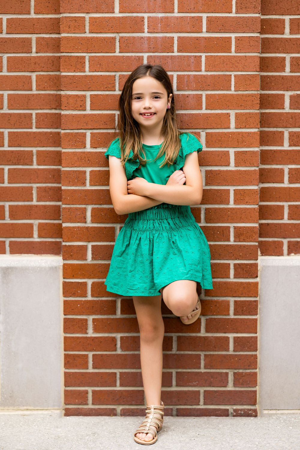 Child in emerald green dress posing playfully on brick wall ledge.