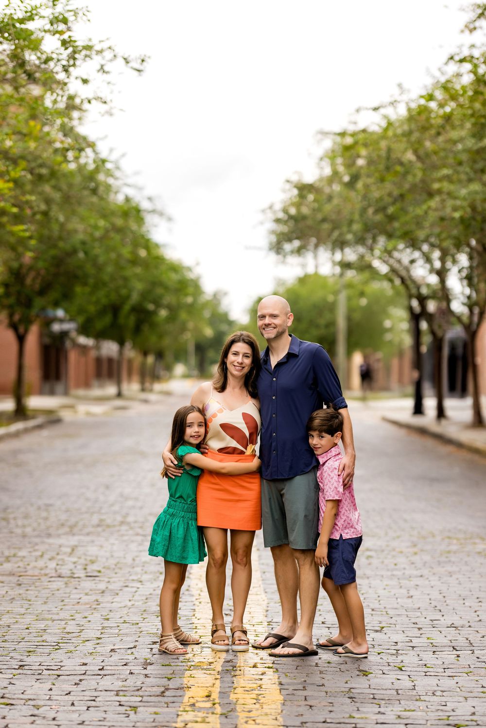 A family stands together on a cobblestone street lined with trees in an urban setting.