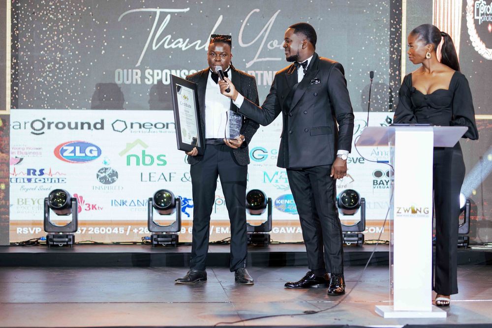 People in formal attire interact on stage at an event with sponsor logos and moving lights visible in the background.