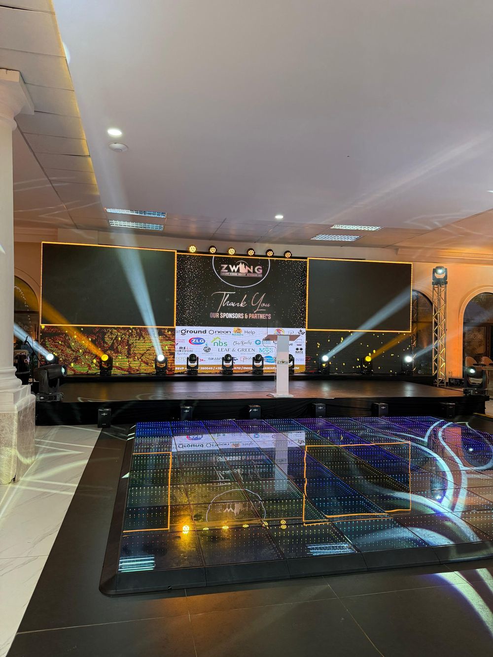 Modern event venue with illuminated stage and dramatic lighting featuring dark backdrop and reflective glass paneling.