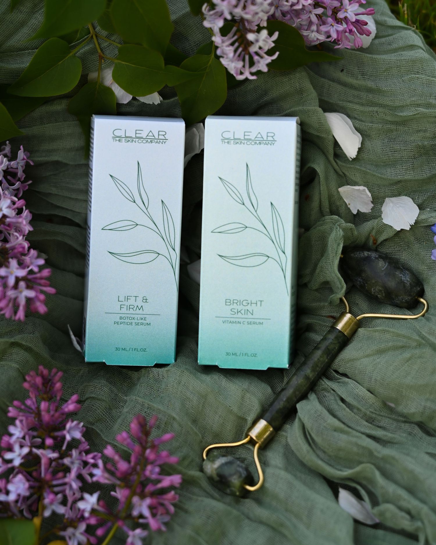 Clear Skincare Lift and Firm and Bright Skin serum boxes with a jade roller among lilac blooms on a leaf.