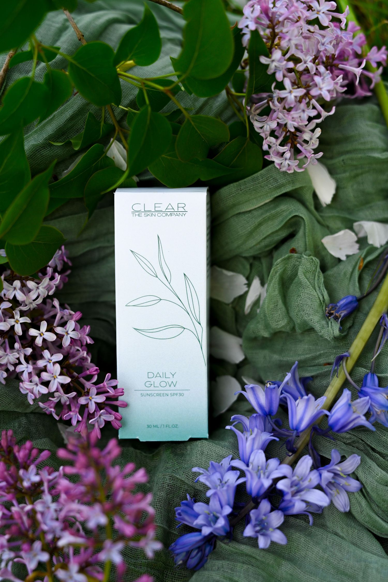 CLEAR The Skin Company Daily Glow facial radiance spray box surrounded by purple lilac and blue flowers.