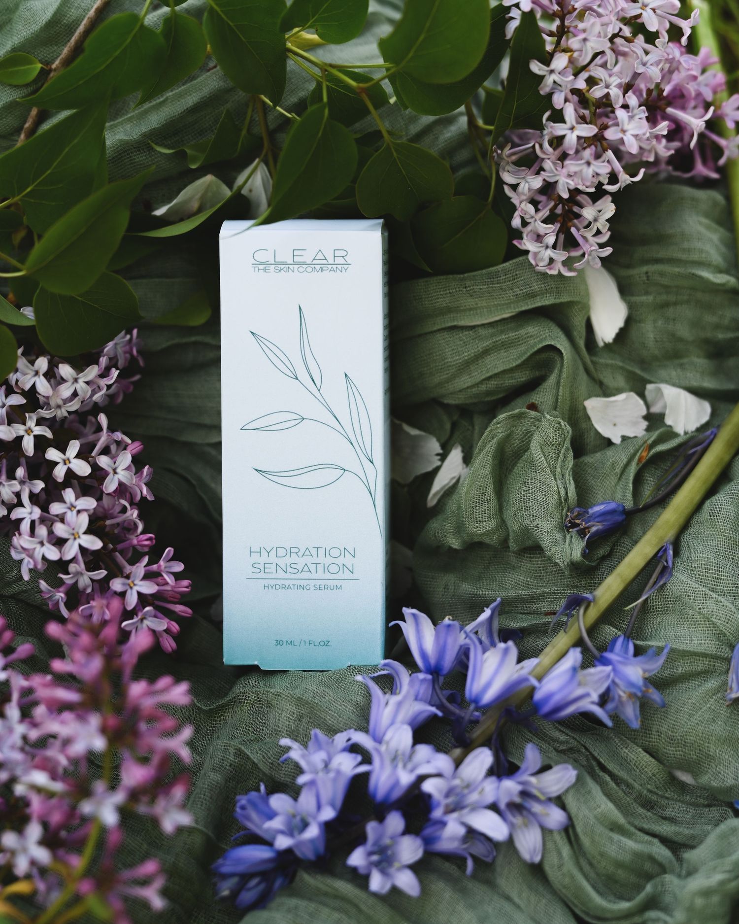 Clear Skincare Hydration Sensation serum box surrounded by purple lilac and blue flowers on green fabric.
